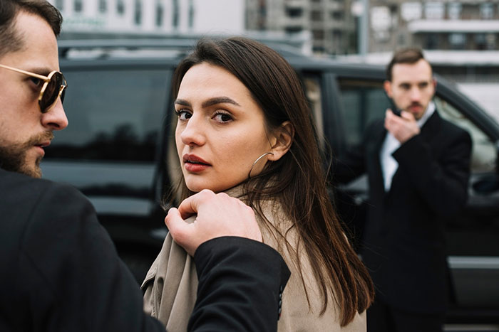 Woman looking back as she stands near a man in a suit, with another man in the background near a black car.