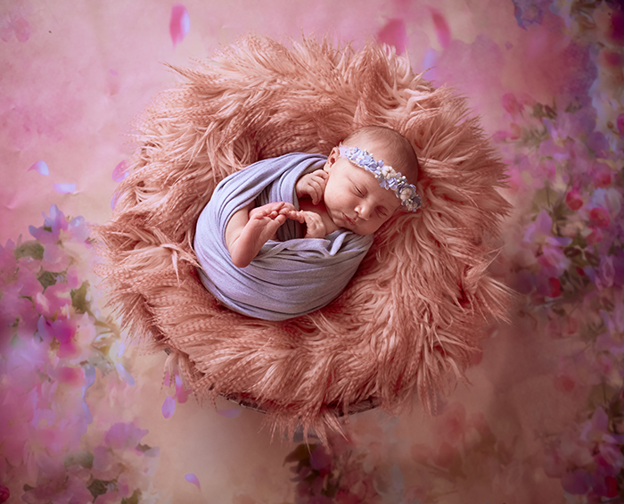 Newborn sleeping in a pink furry nest, wrapped in a purple swaddle with a floral headband.