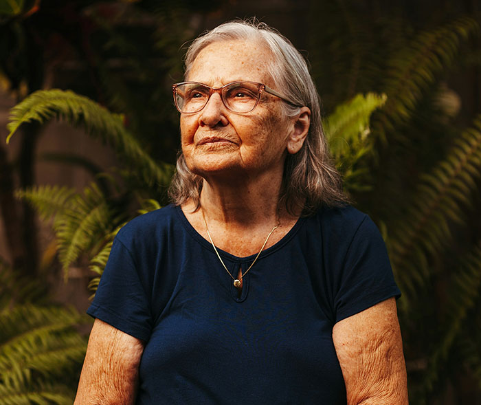 Elderly woman in glasses looking thoughtful, representing suspecting fraud and debt issues in a garden setting.