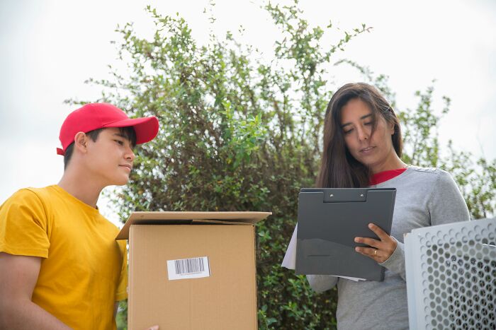 A delivery person in a red cap hands a package to a woman writing on a clipboard.