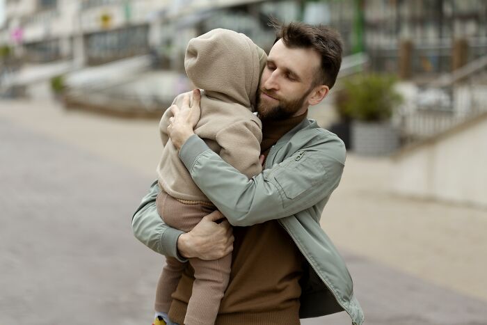 A man in a green jacket warmly embraces a child in a brown hoodie on the street.