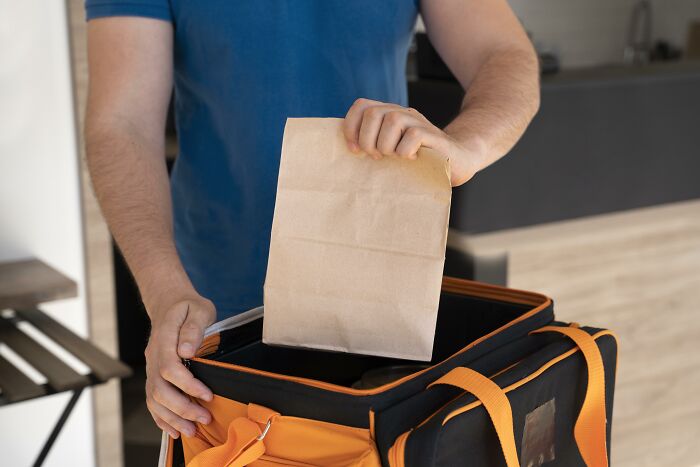 A delivery person in a blue shirt places a paper bag into an orange insulated bag, highlighting a strange delivery scenario.