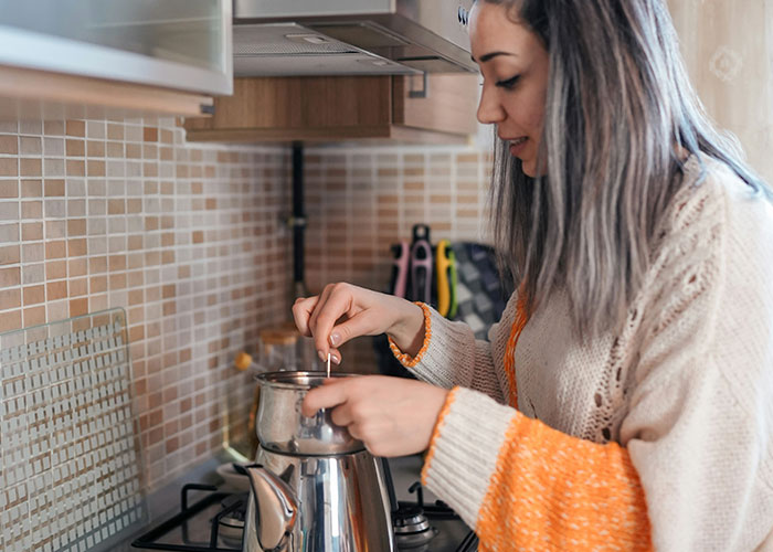 Woman in cozy sweater making tea on stove in kitchen, illustrating times police were called for dumbest reasons.