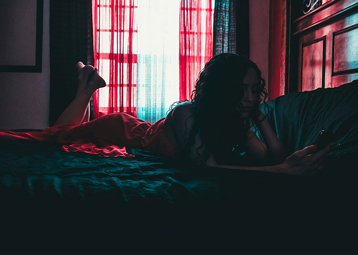 Woman lying on a bed in a dimly lit room, using her phone near red curtains, illustrating police called for dumb reasons.