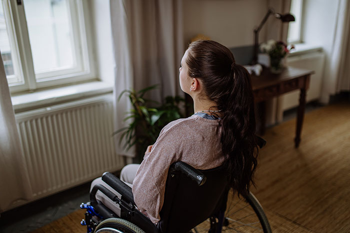 Teen girl in a wheelchair looking out a window, reflecting on family dynamics and responsibilities.