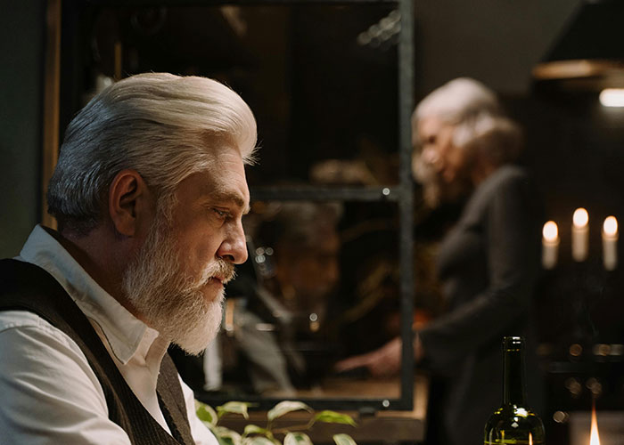 Elderly man with white hair at Thanksgiving dinner, looking thoughtful while a woman stands in the background.