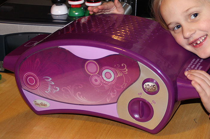 Child smiles beside an Easy-Bake Oven, showcasing a popular item among things people bought as adults denied in childhood.
