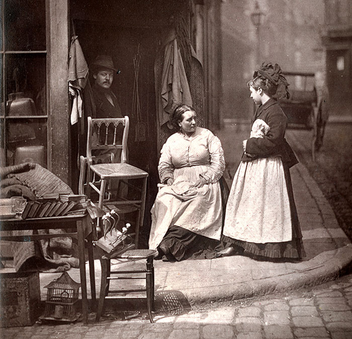 Victorian street scene with women and a man gathered outside a workshop, depicting a romanticized past.
