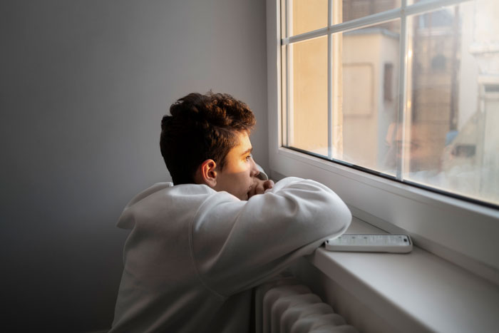 Young man in a white hoodie gazing thoughtfully out the window, with a smartphone beside him. Young man in a white hoodie gazing thoughtfully out the window, with a smartphone beside him.