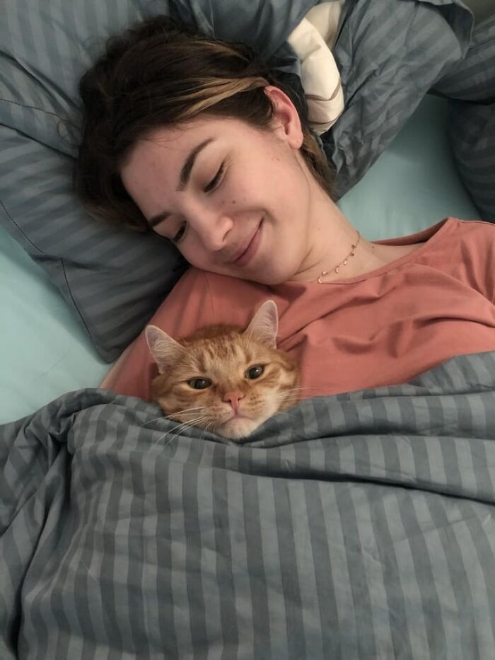 Woman and orange tabby cat catnapping cozily together under gray striped blankets on a bed.