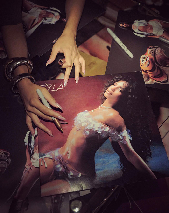 A close-up of hands signing posters featuring Tyla in a glamorous outfit made of translucent sequins. The scene includes markers, bold jewelry, and multiple copies of the poster laid out on a table, showcasing her artistic persona.