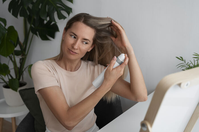 Woman using dry shampoo, applying it to her hair in front of a mirror, illustrating hygiene hacks.