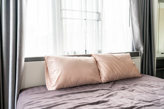 Bed with pink pillows and grey sheets next to a window, illustrating hygiene hacks. 