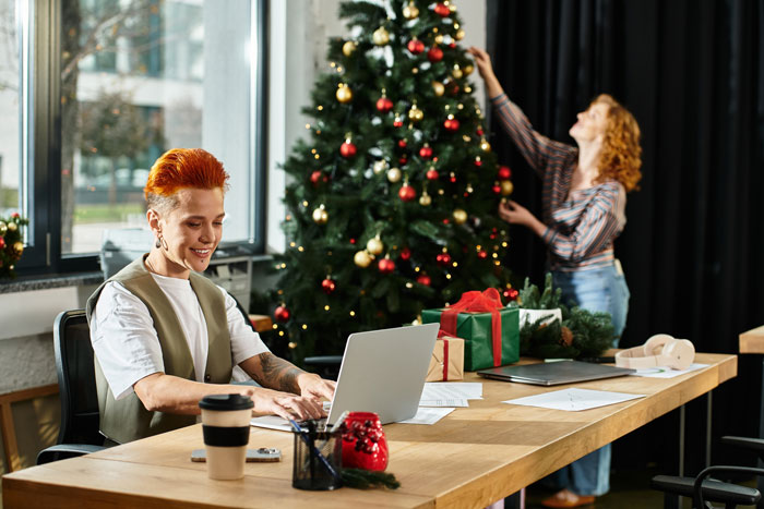 Woman decorating Christmas tree, focusing on Secret Santa planning, with a man using a laptop in the office.