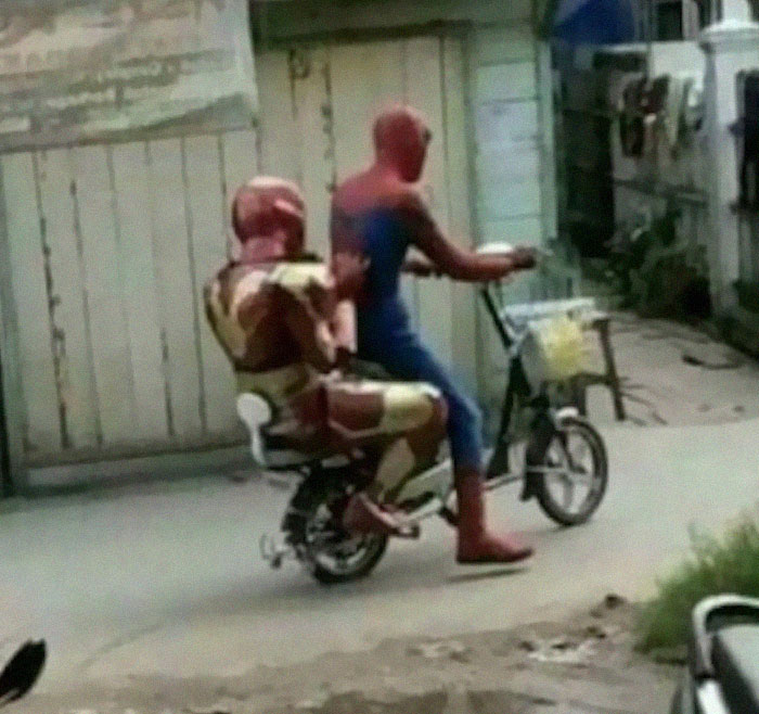 Spider-Man and Iron Man costumed figures ride a bicycle together in a humorous street scene.