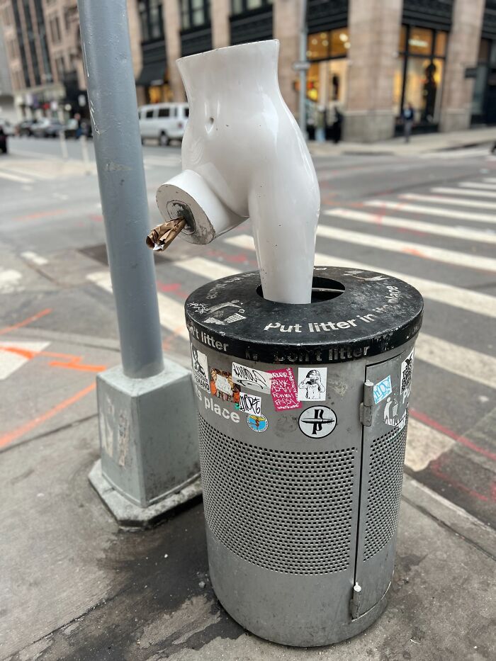 Bizarre street art of a torso sculpture on a trash can, creating a humorous scene on a city sidewalk.