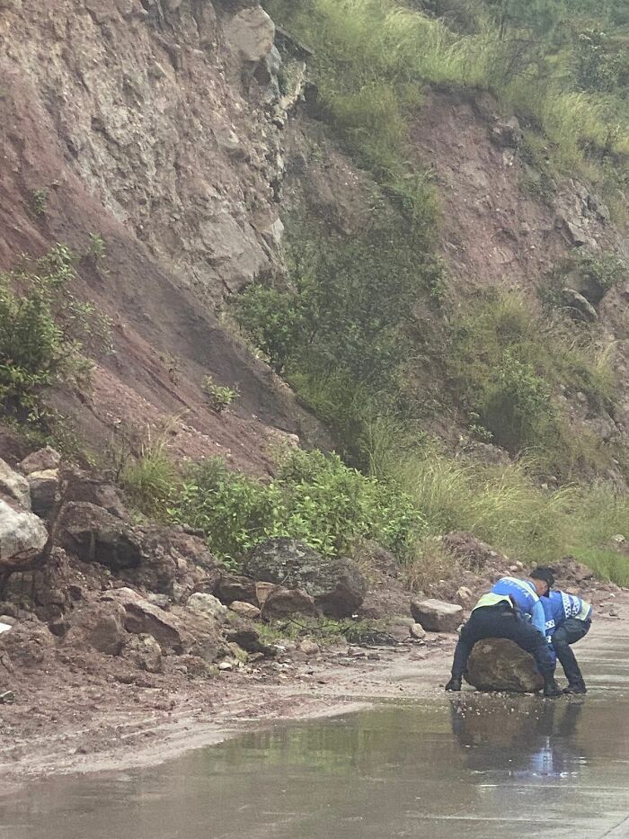 Two people in uniform roll a large rock on a muddy roadside, showcasing a bizarre scene.