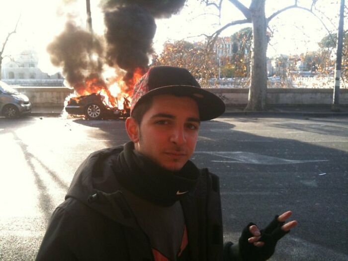 Person standing near a burning car in a bizarre street scene.