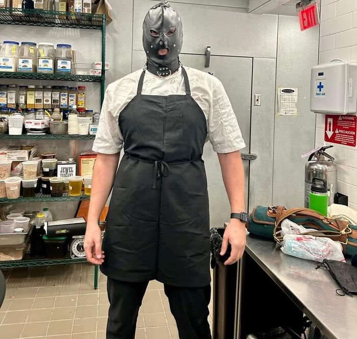 A person in a kitchen wearing a black apron and an unusual mask, embodying bizarre social media humor.