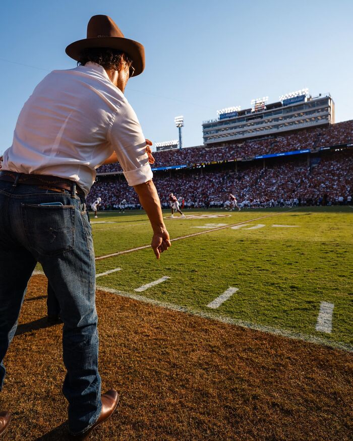 Man in a hat gestures on a football field during a game, with a full stadium in the background.
