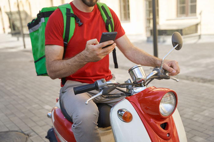 DoorDash driver on a scooter checks phone, sporting a red shirt and green backpack on a city street.