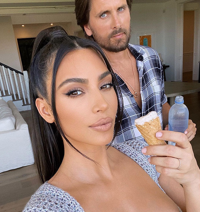 Kim Kardashian holding an ice cream cone while posing for a selfie in a cozy indoor setting, with a man in a plaid shirt holding a water bottle in the background.