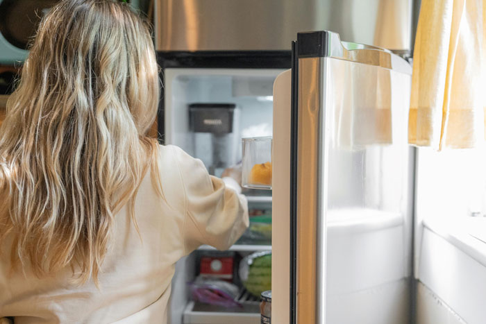 Woman with long hair opens fridge, reaching for food, in well-lit kitchen setting.