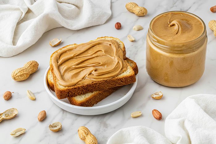 Jar of peanut butter and peanut butter toast on a table, surrounded by peanuts, highlighting allergy discussion context.