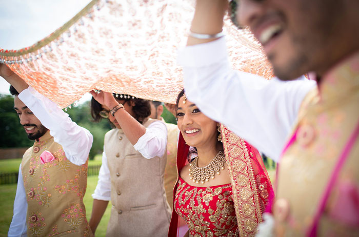 Bride in traditional attire smiling joyfully under ornate canopy at an outdoor wedding ceremony, surrounded by guests.