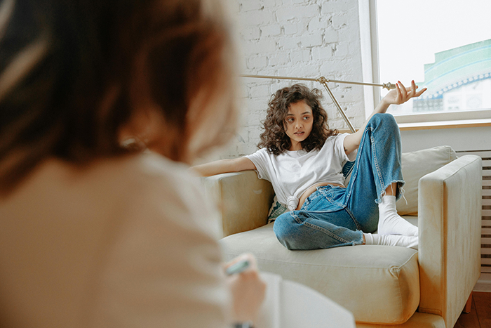 Woman facing poor treatment from MIL, sitting on a couch in casual clothes, gesturing in frustration.