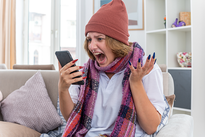 Woman in beanie and scarf looking surprised while using a smartphone on a couch, indoors.