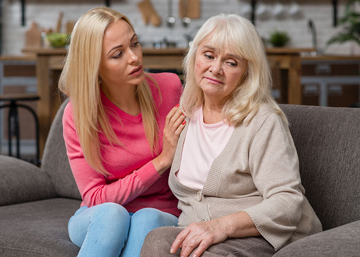 Daughter comforting concerned mother-in-law on a couch, expressing support and empathy.