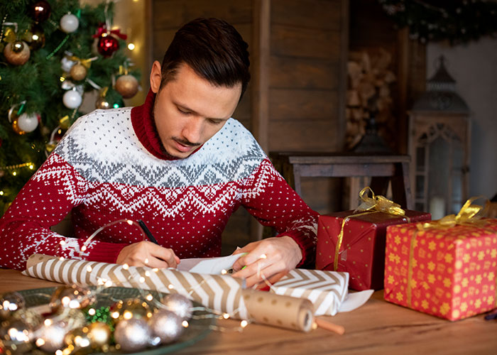 Man wrapping Christmas gifts at a wooden table, surrounded by festive decorations, focusing on holiday preparations.