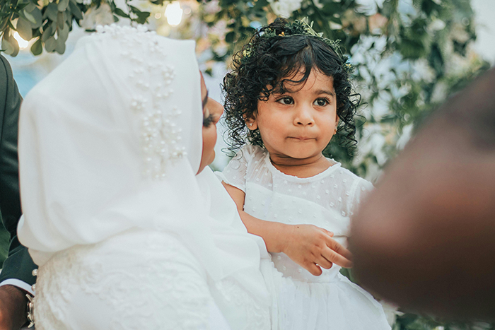 Child at a wedding, surrounded by greenery, holding onto an adult, illustrating family dynamics at events.
