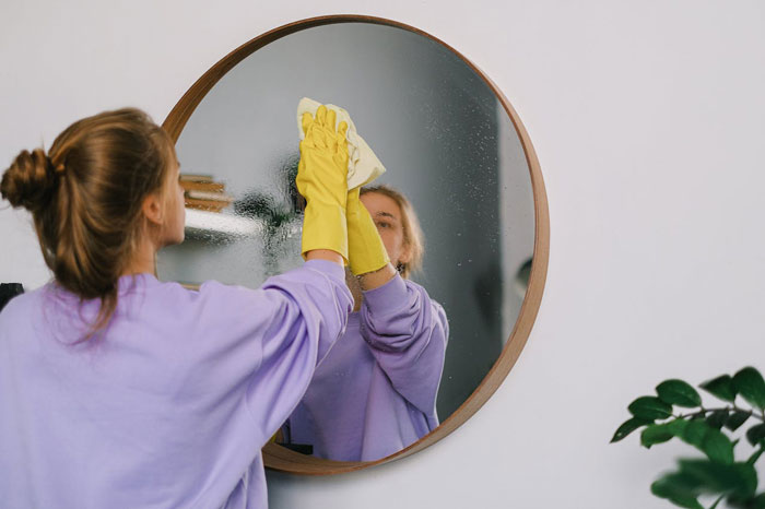 Woman in yellow gloves cleaning a mirror, reflecting a tidy room.