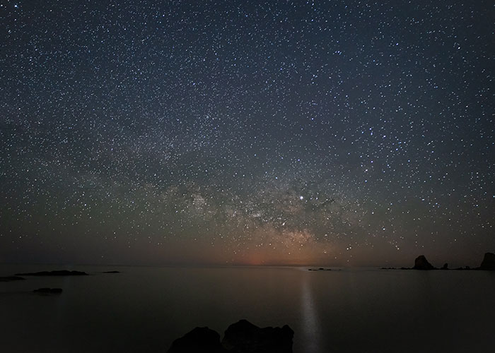Starry night sky over calm sea with silhouetted rocks, creating a mesmerizing and tranquil ocean scene.