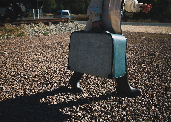 A person in boots walks on gravel carrying a vintage suitcase, outdoors.