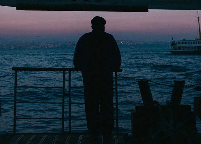 Silhouette of a person on a ship at sea, with a distant city skyline, creating a cautious mood.