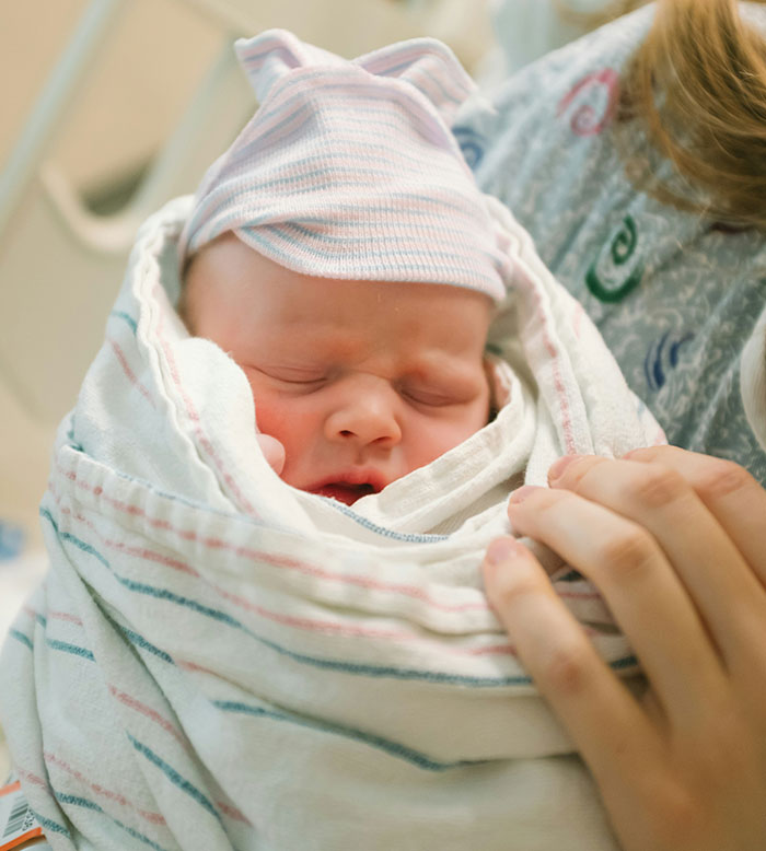 Newborn baby wrapped in a striped blanket and hat, held by a maternity nurse.