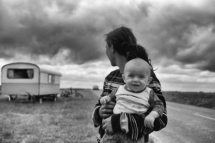 Woman holding baby near trailer under cloudy sky, highlighting maternity challenges.