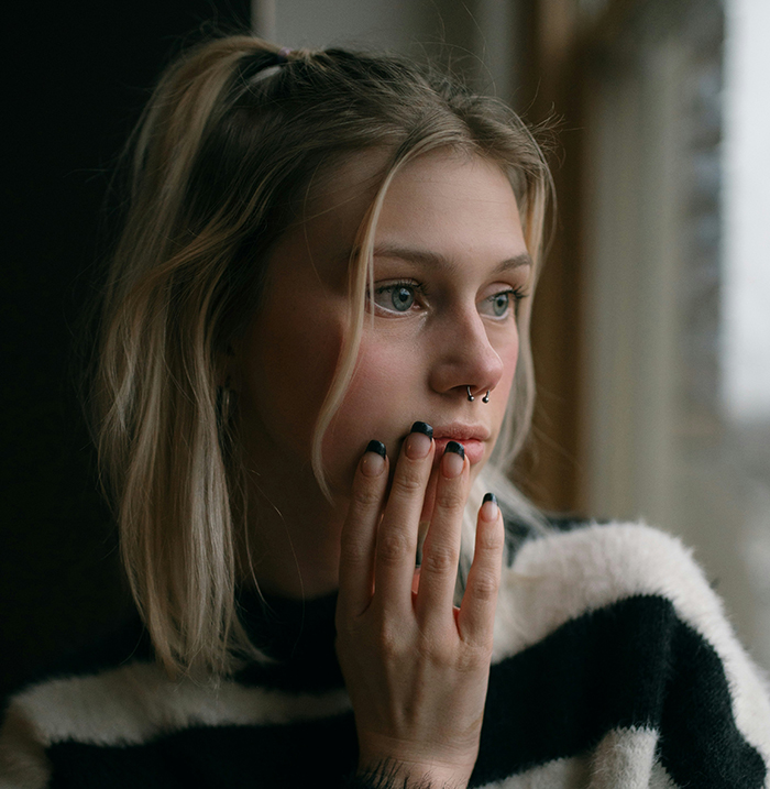 Young woman gazing thoughtfully out a window, hand on chin, reflecting on maternity stories in a cozy setting.