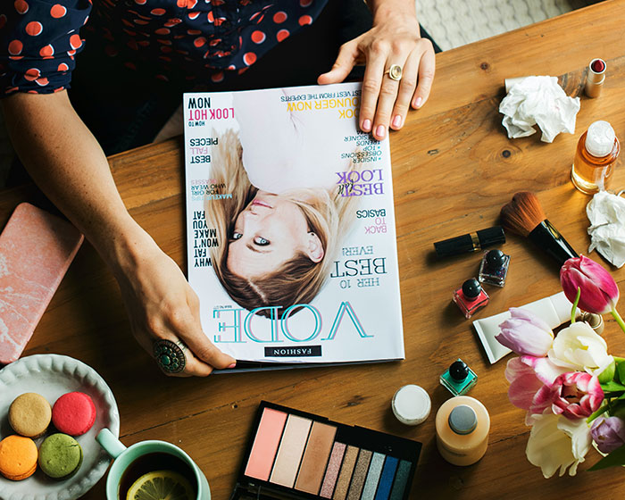 A person holds a fashion magazine surrounded by makeup and flowers, suggesting a Secret Santa gift idea.