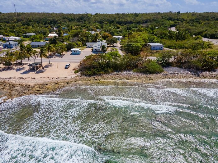 Aerial view of a coastal village with green trees and small houses, waves crashing on the beach, worst city country trip.