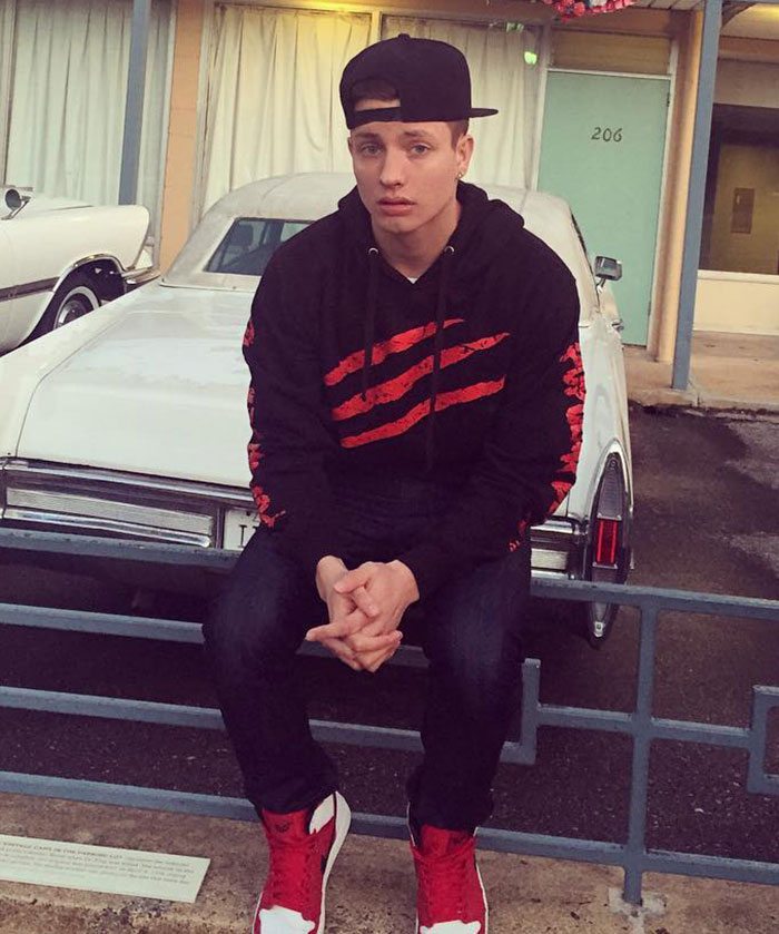 Young Matt Rife in a black hoodie and red shoes sitting on a fence in front of a vintage car