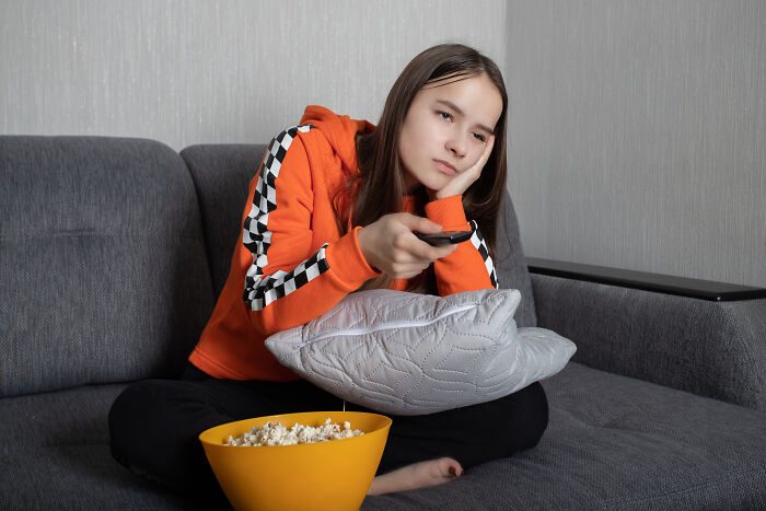 Rich kid on a gray couch in a bright orange hoodie, holding a remote, bored, with popcorn in a yellow bowl.