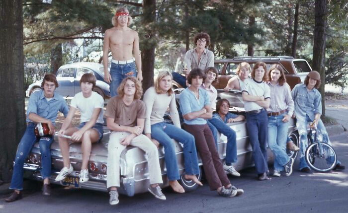 Teenagers in '70s high school attire, gathered around a car, showcasing retro fashion and hairstyles.