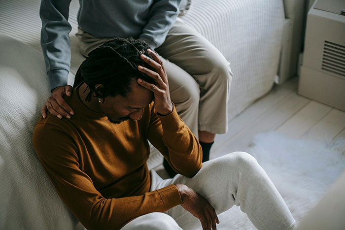 A worried man in a brown sweater sits on the floor, comforted by a person beside him, symbolizing tension over a birthday incident.