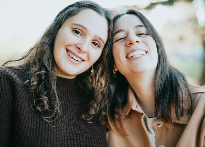 Two smiling women outdoors; one wears a brown sweater, the other a beige coat, relating to a wedding speech incident. Two smiling women outdoors; one wears a brown sweater, the other a beige coat, relating to a wedding speech incident.
