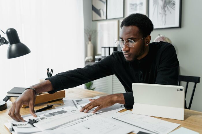 Man in glasses organizing financial documents at a desk, surrounded by papers and a tablet.