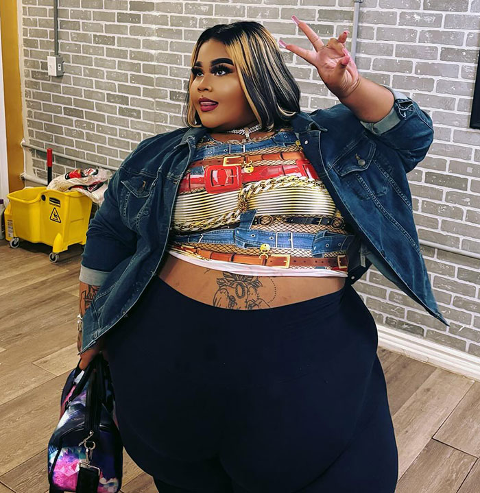 Plus size woman in colorful top and denim jacket poses with a peace sign in a brick-walled room.